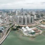 An aerial view of Johor Bahru city, featuring modern skyscrapers, a waterfront, highways, and ongoing construction, highlighting the city’s potential for homestay management opportunities.