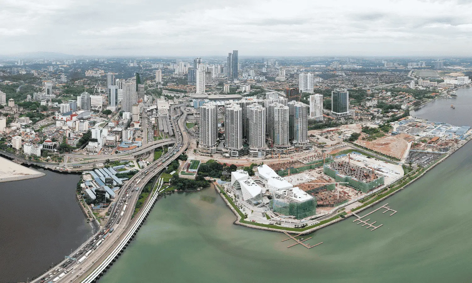 An aerial view of Johor Bahru city, featuring modern skyscrapers, a waterfront, highways, and ongoing construction, highlighting the city’s potential for homestay management opportunities.