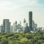 A cityscape of Bangkok, Thailand, featuring modern high-rise buildings and skyscrapers with lush green trees in the foreground under a clear sky
