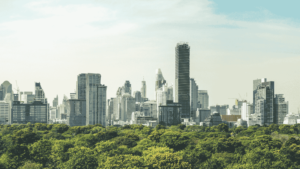 A cityscape of Bangkok, Thailand, featuring modern high-rise buildings and skyscrapers with lush green trees in the foreground under a clear sky