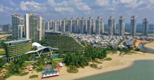 Aerial view of Forest City in Johor, Malaysia, showcasing modern high-rise condominiums, uniquely designed green-roofed buildings, and neatly arranged residential villas along a sandy beachfront, with lush greenery and clear skies in the background.