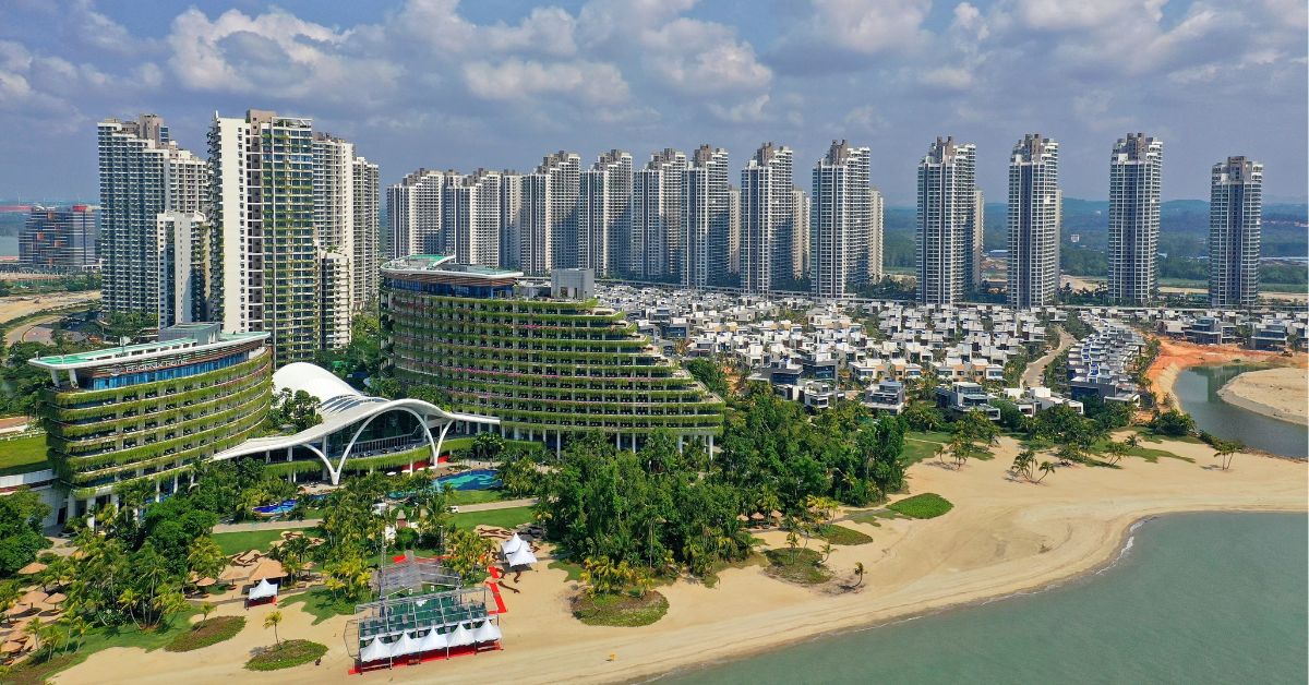 Aerial view of Forest City in Johor, Malaysia, showcasing modern high-rise condominiums, uniquely designed green-roofed buildings, and neatly arranged residential villas along a sandy beachfront, with lush greenery and clear skies in the background.